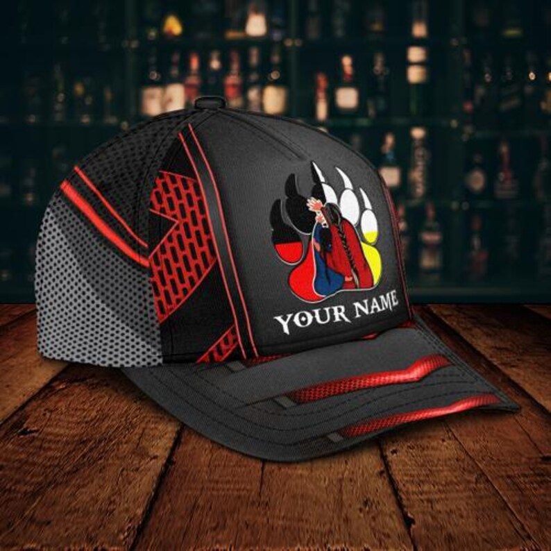 May include: Black baseball cap with a mesh back and red accents. The cap features a graphic of a Native American figure within a bear paw print, with the text "YOUR NAME" below the graphic. The cap is on a wooden surface.