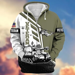 Pode incluir: Um casaco com capuz e z&iacute;per com tema militar. O design apresenta um tanque, helic&oacute;pteros e a palavra "VETERAN" em branco. A manga esquerda tem uma bandeira americana. O esquema de cores inclui verde-oliva, branco e preto.