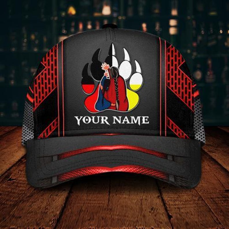 May include: Black baseball cap with a Native American design. The cap features a bear paw print with a figure in traditional clothing, and the text "YOUR NAME". Red and black geometric patterns adorn the sides.