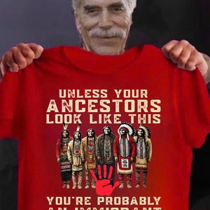 May include: Red t-shirt with the text "UNLESS YOUR ANCESTORS LOOK LIKE THIS YOU'RE PROBABLY AN IMMIGRANT". The shirt features a graphic of six Native American figures and a red handprint.