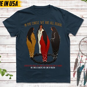Puede incluir: Camiseta azul marino con el texto "IN THE CIRCLE WE ARE ALL EQUAL". La camiseta presenta una ilustraci&oacute;n de tres figuras en un c&iacute;rculo. Las palabras "NO ONE IS ABOVE, NO ONE IS BELOW" tambi&eacute;n est&aacute;n impresas. Hecho en EE. UU.