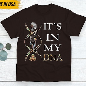 May include: A dark brown t-shirt with the text "IT'S IN MY DNA" in white. The design includes a DNA strand with Native American-themed illustrations. The shirt is labeled "MADE IN USA" in a yellow box.
