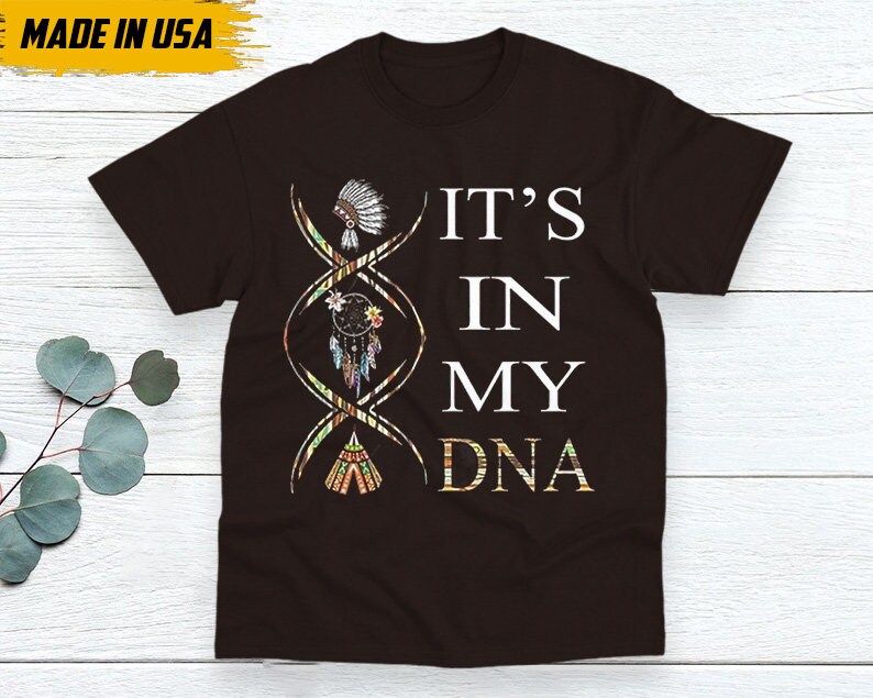 May include: A dark brown t-shirt with the text "IT'S IN MY DNA" in white. The design includes a DNA strand with Native American-themed illustrations. The shirt is labeled "MADE IN USA" in a yellow box.