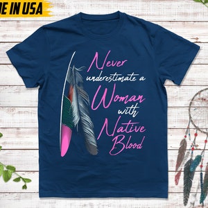 May include: Navy blue t-shirt with the text "Never underestimate a Woman with Native Blood" in pink script. The shirt features a feather graphic with pink, green, and gray accents. The words "Made in USA" are in the top left corner.