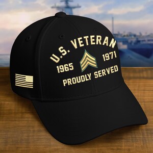 May include: Black baseball cap with gold embroidered text and an American flag patch. The text reads "U.S. Veteran 1965-1971 Proudly Served" with a military insignia. The cap is on a wooden surface.