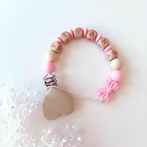 May include: A pacifier clip featuring a circular arrangement of light pink and beige silicone beads, interspersed with wooden cube beads spelling the name ELIANNA. A metal clip is attached to a heart-shaped beige silicone piece and a pink bow.