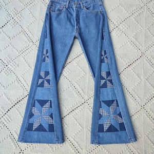 Peut inclure: Jean bleu clair avec jambes évasées. Les jeans présentent des motifs patchwork dans les tons de bleu sur les coutures extérieures. Le patchwork comprend des motifs d'étoiles géométriques. Le jean est posé à plat sur une surface crochetée blanche.