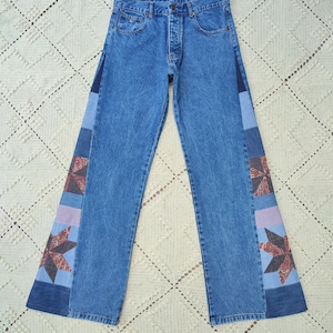 May include: Light blue denim jeans with patchwork star designs on the outer seams. The jeans have a button fly and classic five-pocket styling. The patchwork features shades of blue, red, and purple.