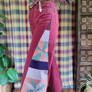 Op de afbeelding: Bordeauxrode broek met patchwork detail. Het patchwork heeft een sterrenpatroon met groene bloemenstof, een marineblauwe streep en een lichtblauw-wit geruit patroon. De broek heeft een wijde pijp en een hoge taille.