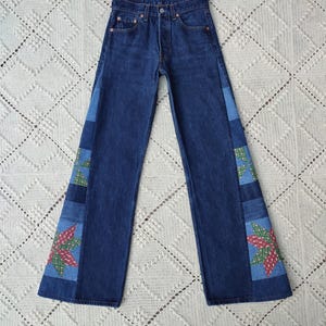 Könnte beinhalten: Dunkelblaue Jeans mit Patchwork-Akzenten. Die Jeans haben ein klassisches Fünf-Taschen-Design und einen Knopfverschluss. Das Patchwork umfasst blaue und grüne geometrische Muster sowie rot-grüne Blumenmuster.