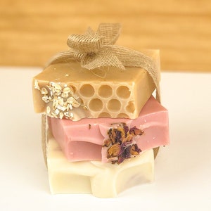 May include: Three bars of handmade soap stacked on top of each other. The top bar is a light brown color with a honeycomb pattern and oatmeal flakes. The middle bar is a light pink color with dried rose petals. The bottom bar is a white color. The bars are tied together with a piece of twine.
