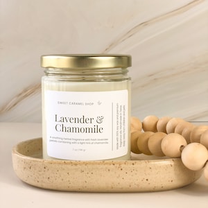 May include: A glass jar candle with a gold lid, labeled "Lavender & Chamomile" with the text "Sweet Caramel Shop" and "7 oz 198 gr". The candle is sitting on a light brown oval ceramic dish with a wooden bead garland in the background.
