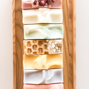 May include: A wooden tray with a variety of handmade soap bars. The bars are arranged in a stack and include colors like pink, green, yellow, and white. Some bars have decorative elements like dried flowers and oatmeal.