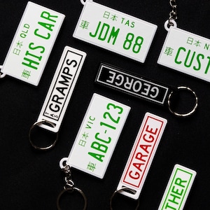 May include: A collection of seven keychains with different designs. The keychains are white, black, and red with green text. The text on the keychains includes "HIS CAR", "GRAMPS", "ABC-123", "GARAGE", "THER", "GEORGE", "JDM 88", and "CUSTOM".
