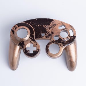 May include: A video game controller faceplate featuring a marbled pattern of metallic bronze and deep black. The shell includes cutouts for the analogue sticks, directional pad, and action buttons, showcasing a smooth, glossy finish throughout.