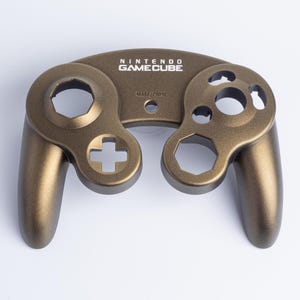 May include: A metallic bronze-coloured replacement front shell for a Nintendo GameCube controller. The shell features the Nintendo GameCube logo printed in white at the top centre and the word START/PAUSE embossed below the logo area.