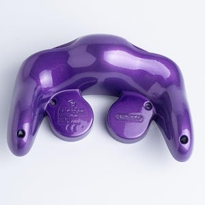 May include: A metallic purple Nintendo GameCube controller shown from the back. The casing features a glossy finish with visible screw holes and moulded text including the Nintendo logo, model number DOL-003, and the text Made in China.