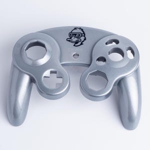 May include: A silver-coloured game controller faceplate featuring a black graphic of a duck wearing goggles centred at the top. The shell has cutouts for buttons, joysticks, and a directional pad, with a smooth, metallic finish throughout the surface.