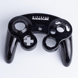 May include: A black Nintendo GameCube controller front shell featuring a glittery finish. The plastic housing includes cutouts for the analogue sticks, buttons, and D-pad, with the Nintendo GameCube logo printed in white text at the top centre.