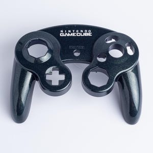 May include: A black, glittery Nintendo GameCube controller front shell casing. The plastic housing features cutouts for the analogue sticks, D-pad, and action buttons, with the Nintendo GameCube logo printed in white text at the top centre.