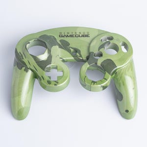 May include: A Nintendo GameCube controller shell featuring a glossy olive green and dark green camouflage pattern. The front face displays the Nintendo GameCube logo in black text, with cutouts for buttons, joysticks, and the directional pad.