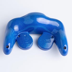 May include: A glossy, metallic blue game controller shell with a glittery finish. The piece features two downward-curving handles, a central bridge, and two circular cutouts for analog sticks, with visible screw holes and moulded Nintendo branding.