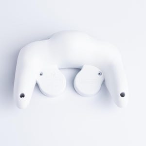 May include: A solid white video game controller shown from the back side. The device features two ergonomic hand grips, a central curved body, and two circular protrusions on the rear panel with visible screw holes and embossed branding text.