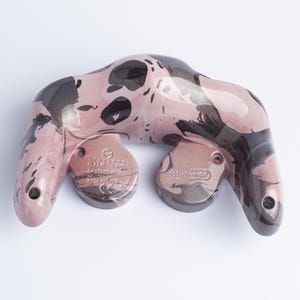 May include: A video game controller back shell featuring a glossy, marbled pattern of dusty pink and dark charcoal black. The moulded plastic surface displays the embossed Nintendo logo and regulatory text, including the model number DOL-003.