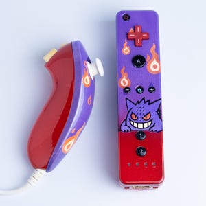 May include: A custom video game controller and nunchuk set featuring a purple and red gradient finish. The controller displays a graphic of a purple ghost character with a wide grin, surrounded by orange flame icons, black buttons, and a red D-pad.