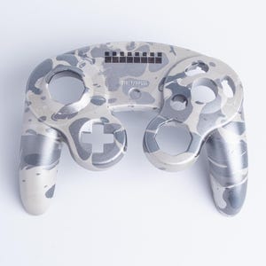 May include: A game controller shell featuring a grey and white camouflage pattern. The front face includes cutouts for buttons and joysticks, with the text START/PAUSE printed in the centre above a small circular opening and a rectangular grid.
