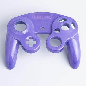 May include: A purple Nintendo GameCube controller front shell casing with a glossy, metallic finish. The plastic faceplate features cutouts for the analogue sticks, D-pad, and action buttons, with the Nintendo logo embossed at the top centre.