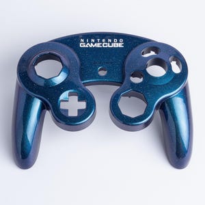 May include: A dark blue, glitter-finish replacement front shell for a Nintendo GameCube controller. The shell features the white Nintendo GameCube logo printed at the top centre, with cutouts for the analogue sticks, buttons, and D-pad.