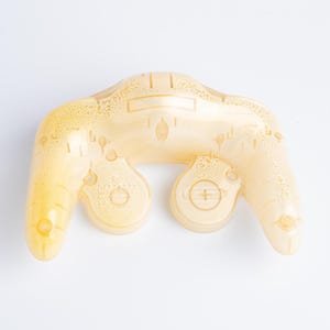 May include: A translucent, pale yellow game controller shell featuring a moulded ergonomic design with two downward-curving grips, a central rectangular cutout, and circular button housings. The surface has a textured, bubbly appearance.