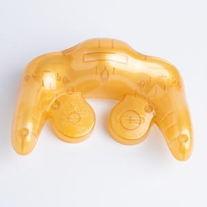 May include: A translucent amber-coloured video game controller shell featuring a smooth, curved design. The plastic casing displays internal structural details and moulded circular ports, showcasing a semi-transparent finish that reveals the inner components.