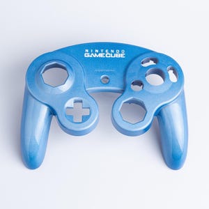 May include: A metallic blue Nintendo GameCube controller front shell casing. The plastic faceplate features cutouts for the analogue sticks, D-pad, and action buttons, with the white Nintendo GameCube logo printed clearly at the top centre.