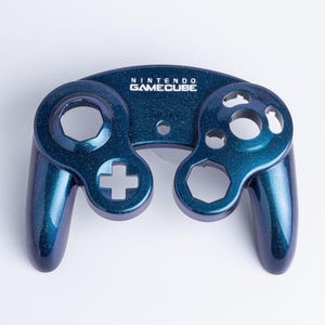 May include: A replacement front shell for a Nintendo GameCube controller featuring a deep metallic blue finish with subtle glitter accents. The shell includes the white Nintendo GameCube logo printed above the central joystick opening.