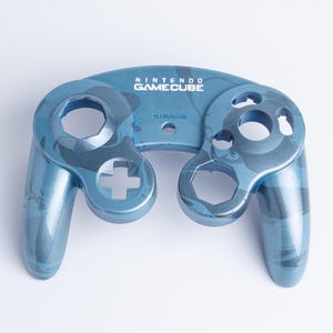 May include: A blue and dark blue camouflage patterned Nintendo GameCube controller front shell replacement. The plastic faceplate features the Nintendo GameCube logo printed in white at the top centre and includes cutouts for buttons.