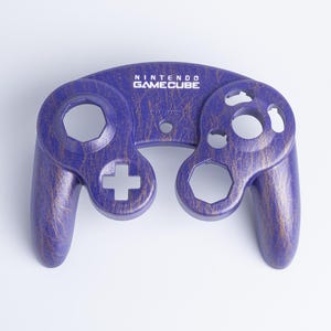 May include: A purple Nintendo GameCube controller front shell featuring a gold-coloured crackle pattern. The shell includes cutouts for the analogue sticks, buttons, and D-pad, with the Nintendo GameCube logo printed in white at the top centre.