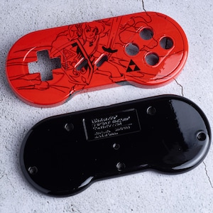 May include: A red Nintendo controller top with a black illustration of a character with a sword and shield. The controller has a directional pad and button holes. A black controller base is below.