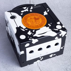 Puede incluir: Una consola Nintendo GameCube negra con un diseño único de estampado de vaca. La consola presenta manchas blancas sobre un fondo negro, con un emblema circular en la parte superior. El emblema tiene un gráfico de vaca y texto.