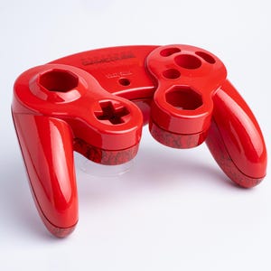 Può includere: Una scocca per controller Nintendo GameCube di colore rosso lucido con una finitura brillante. La parte superiore presenta il logo e la scritta Nintendo GameCube in rilievo. Il corpo ha aperture per i pulsanti e un motivo marmorizzato scuro.