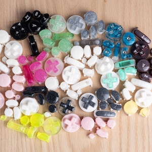 Może przedstawiać: Kolekcja kolorowych, dekoracyjnych przycisków i padów kierunkowych do kontrolerów gier wideo w różnych kształtach i rozmiarach. Przyciski są w odcieniach czerni, bieli, różu, zieleni, błękitu i żółci, rozrzucone na jasnej drewnianej powierzchni.