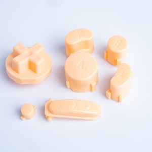 Può includere: Un set di pulsanti in plastica color pesca per controller di gioco, che include una croce direzionale, pulsanti circolari con le lettere A, B, X e Y, un piccolo pulsante rotondo e un pulsante rettangolare lungo, tutti con piccole alette.