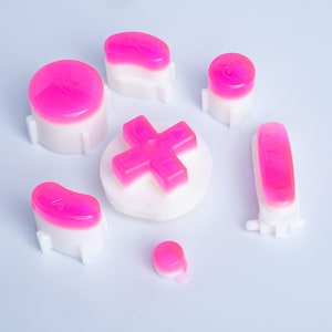 May include: A set of replacement gaming controller buttons featuring a white base and a translucent hot pink top layer. The collection includes a directional pad, various circular and oblong buttons, and a small round button, all with moulded plastic details.
