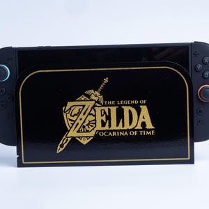 May include: A black gaming console dock featuring a gold-colored The Legend of Zelda Ocarina of Time logo and sword emblem on the front panel. The console has black controllers attached on both sides, with a blue accent on the left joystick.