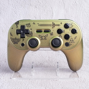 Peut inclure: Une manette de jeu vidéo dorée avec des boutons noirs et un pavé directionnel. La manette est décorée de dessins au trait noirs de divers symboles et motifs. La manette est exposée sur un présentoir en plastique transparent.