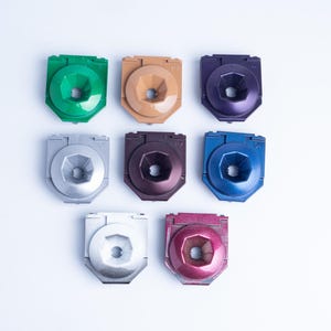 Puede incluir: Una colección de ocho pequeños componentes de plástico de forma cuadrada dispuestos en tres filas. Cada pieza presenta una hendidura hexagonal central y un orificio circular. Los colores incluyen verde, beige, morado, plateado, marrón oscuro, azul, blanco y magenta.