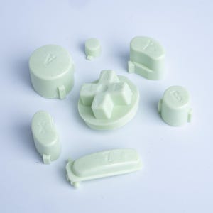 Puede incluir: Un conjunto de botones de repuesto para mando en color verde pálido que incluye una cruceta direccional, botones circulares etiquetados como A, B, X e Y, un botón circular pequeño y un botón rectangular etiquetado como Z, de plástico.