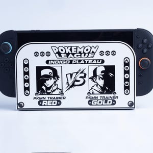 Puede incluir: Una consola de juegos portátil negra con una cubierta de base blanca que presenta un diseño gráfico en blanco y negro. El texto dice POKEMON LEAGUE INDIGO PLATEAU, PKMN TRAINER RED, VS y PKMN TRAINER GOLD, junto a varios iconos elementales.