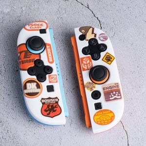 Könnte beinhalten: Zwei weiße und orangefarbene Gamecontroller mit schwarzen Knöpfen und bunten Aufklebern. Ein Controller hat Aufkleber mit "150cc CLASS", "TURBO", "DK BOOSTERS" und "BOWSER motors". Der andere hat Aufkleber mit "ENGINE" und "FIRE MART".